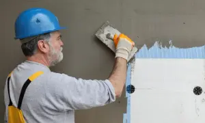 Budowlaniec w niebieskim kasku nakłada zaprawę na styropian. Pękanie kleju na elewacji może wynikać z błędów wykonania.