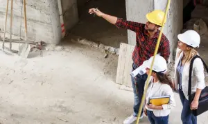 Trzy osoby w kaskach na budowie, mężczyzna wskazuje coś ręką, kobiety trzymają dokumenty.