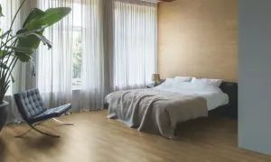 Sypialnia z łóżkiem, fotelem i rośliną. Podłoga z paneli ułożona równolegle do okna, tworząc spójną przestrzeń.