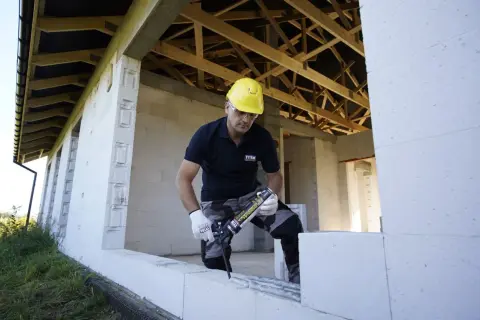 Budowlaniec w żółtym kasku używa pianoklej do murowania, budując dom z białych pustaków.