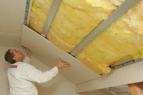 Pracownik montuje płyty gipsowo-kartonowe na poddaszu, nad wełną mineralną. Pokazuje, jak kłaść wełnę na poddaszu.