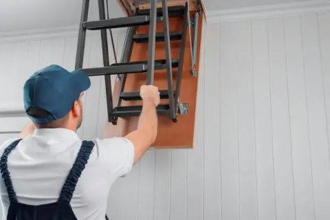 Mężczyzna w czapce i ogrodniczkach rozkłada metalowe schody strychowe.