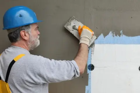 Budowlaniec w niebieskim kasku nakłada zaprawę na styropian. Pękanie kleju na elewacji może wynikać z błędów wykonania.
