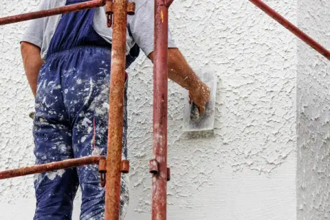 Wykonawca w brudnych ogrodniczkach nakłada tynk na ścianę zewnętrzną, tworząc fakturę baranka.