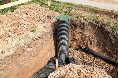 Czarna, karbowana rura drenarska w otulinie, z zieloną pokrywą, umieszczona w wykopie. Wokół żwir i czarna geowłóknina.