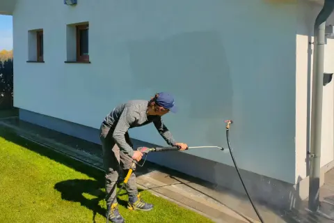 Mężczyzna myjką ciśnieniową czyści elewację z zacieków. Woda rozpryskuje się na białej ścianie domu.