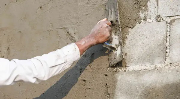 Ręka w białej koszuli tynkuje ścianę z bloczków betonowych kielnią. Proces jak zrobić baranek cementowy w praktyce.