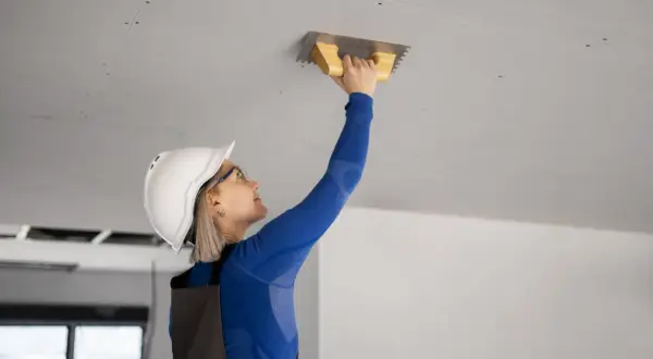 Kobieta w kasku i okularach pracuje przy suficie, używając kielni. To kluczowy etap prac, jak prawidłowo ocieplić strop betonowy.