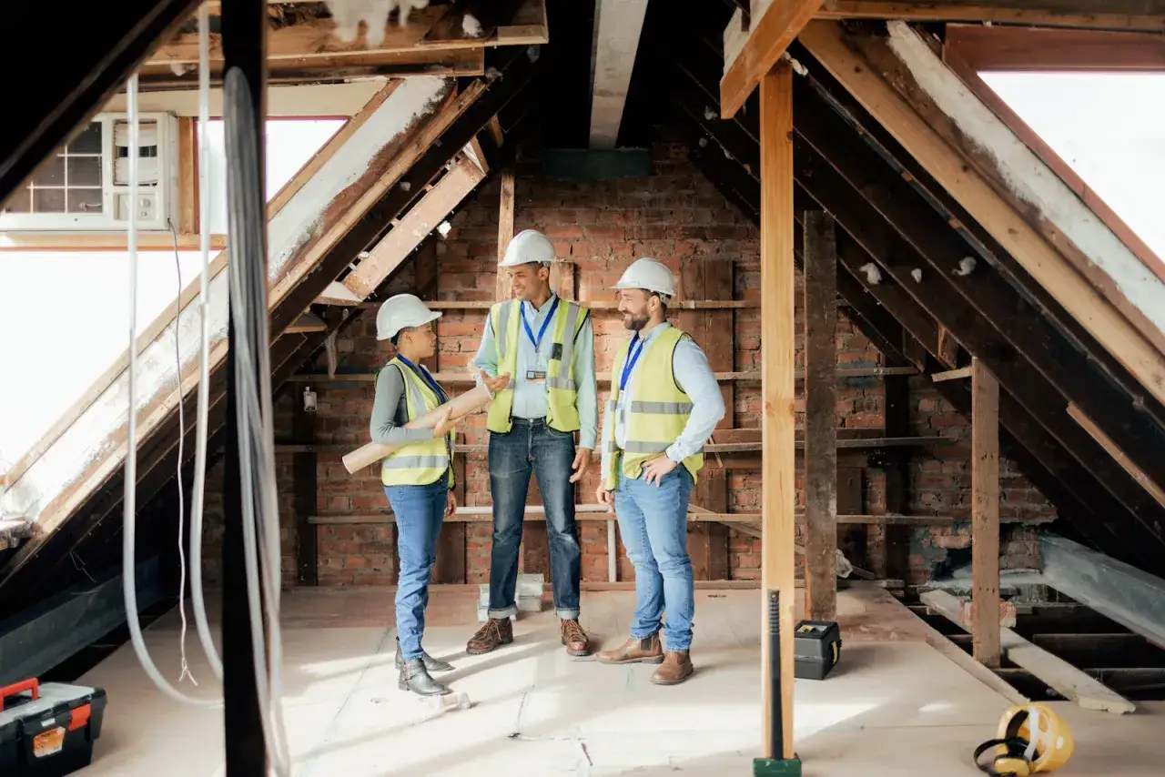 Architekci omawiają, jak zagospodarować poddasze nieużytkowe. Trzy osoby w kaskach i kamizelkach odblaskowych analizują plany w surowym wnętrzu.