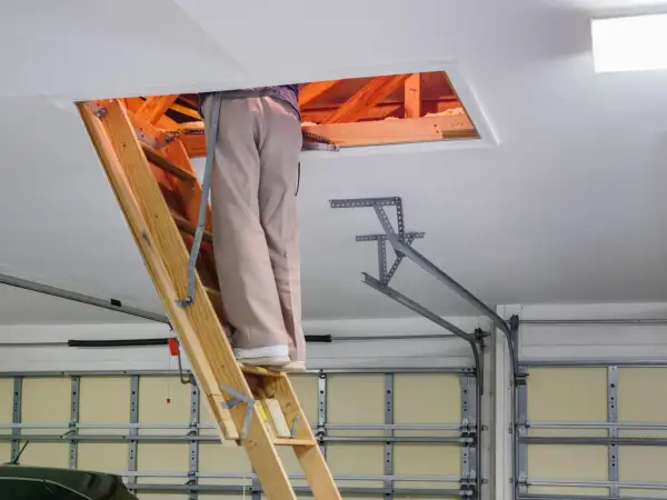 Osoba na drabinie przy wyłazie strychowym. Dowiedz się, jak uszczelnić wyłaz strychowy, by oszczędzać energię.
