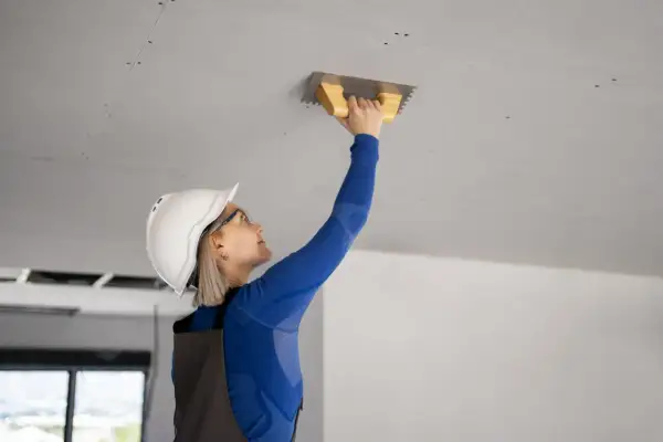 Kobieta w kasku i okularach pracuje przy suficie, używając kielni. To kluczowy etap prac, jak prawidłowo ocieplić strop betonowy.