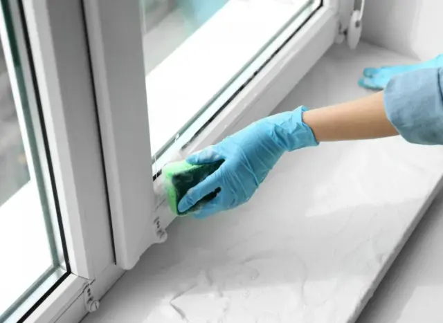 Ręka w niebieskiej rękawiczce czyści parapet gąbką. Dowiedz się, jak usunąć taśmę ze szkła, aby okna lśniły.